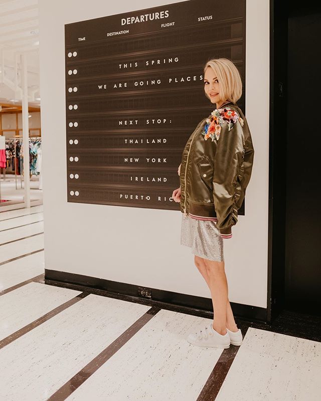 Whitney posing in a floral bomber jacket in front of a departures sign